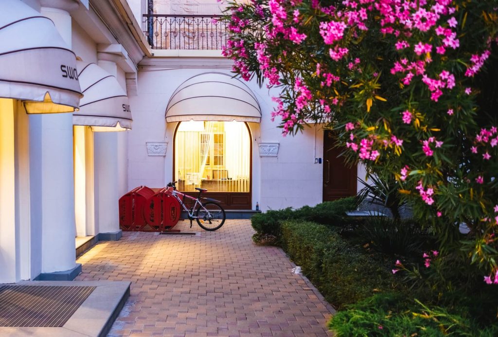 Vista parcial de una construcción elegante con ventanales iluminados y enfrente un parterre de plantas, y flores